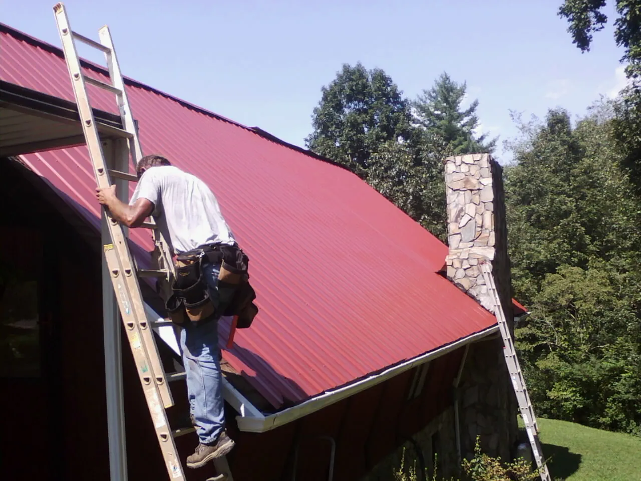 Roofer on a ladder beside a red metal roof and stone chimney — Soffit & Fascia Repair by Roofing Brothers in Hicksville